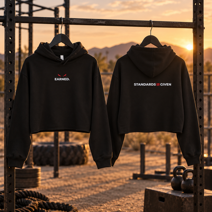 Two black cropped hoodies hanging on a metal rack outdoors at sunset. The front hoodie shows the word 'EARNED.