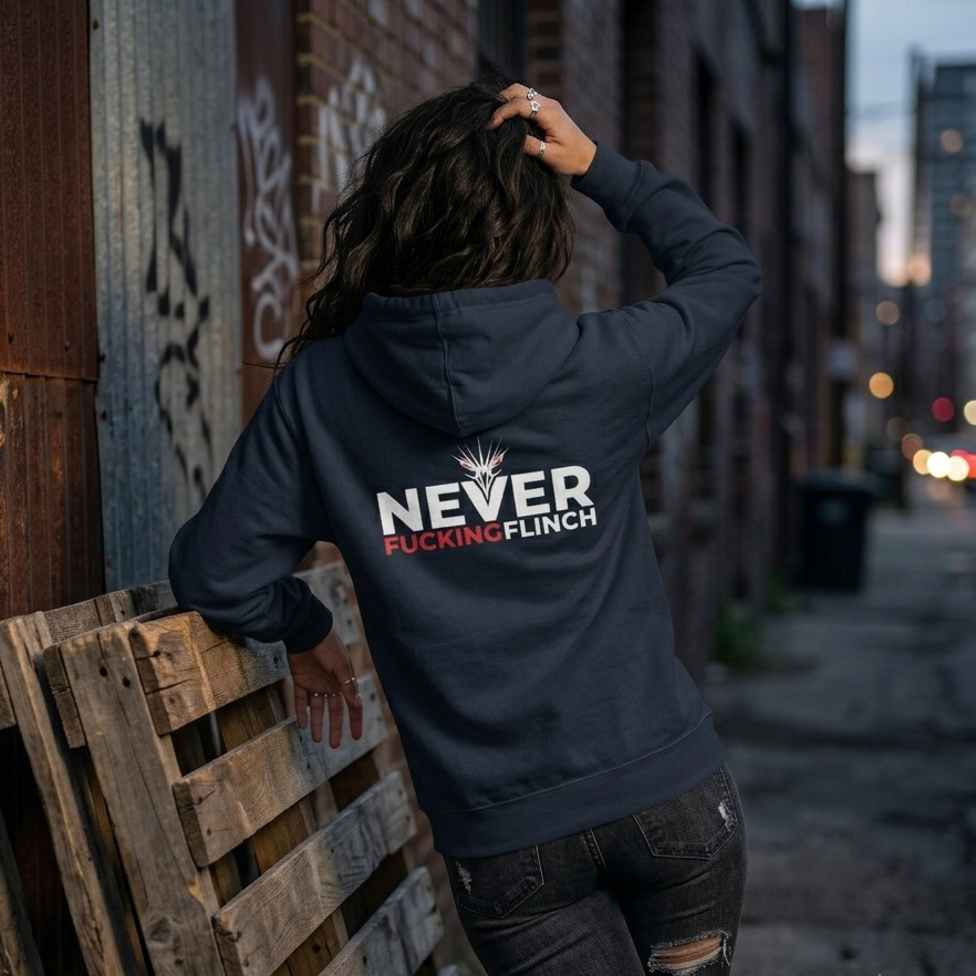 A woman stands with her back to the camera in an urban alley, wearing a navy hoodie with a bold graphic on the back that reads 'NEVER FUCKING FLINCH' in white and red, paired with distressed black jeans.