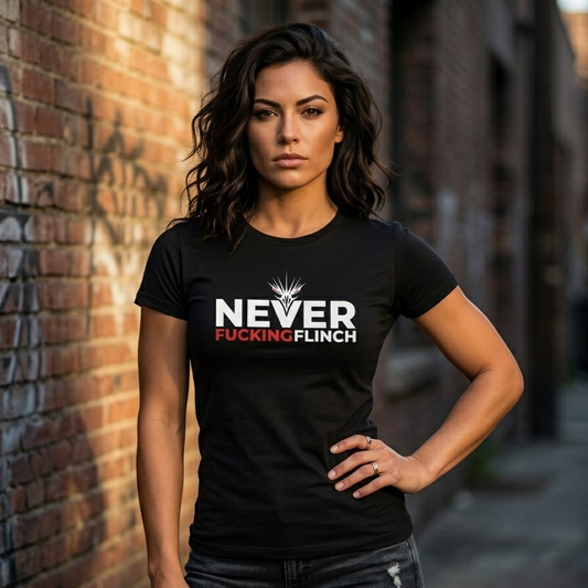 A woman stands in an alley wearing a fitted black T-shirt with a bold white and red graphic reading “NEVER FUCKING FLINCH” on the chest, paired with dark jeans.